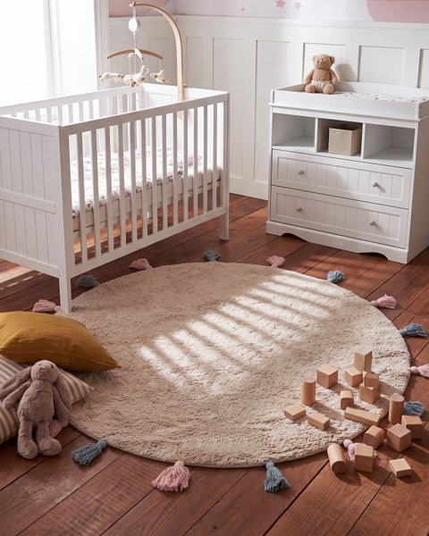 Helles Kinderzimmer mit weißem Gitterbett, Wickelkommode, rundem Teppich mit Quasten und Holzbausteinen auf einem Holzdielenboden.
