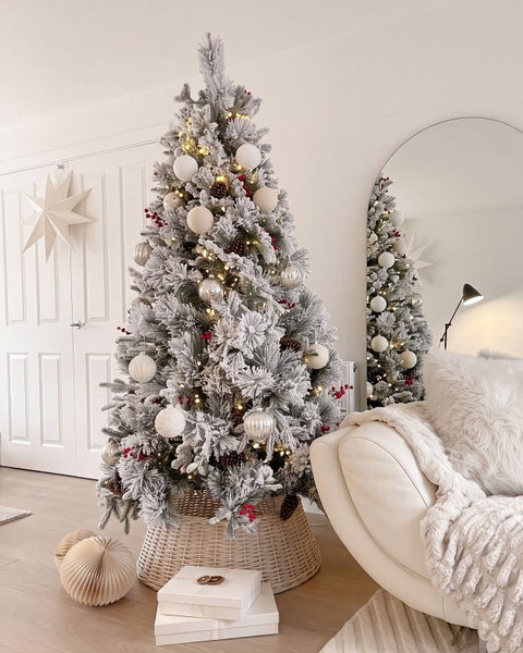 Geschmückter künstlicher Weihnachtsbaum mit Schneeoptik, Lichterkette und Dekoration in einem hellen Wohnzimmer mit Korb als Baumständer.