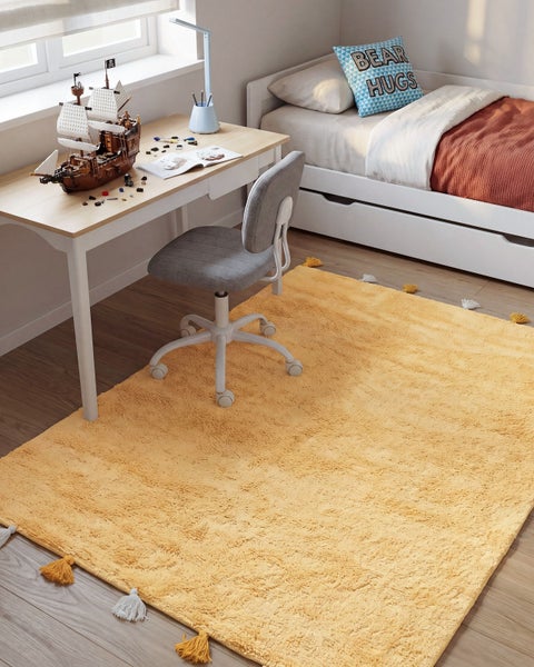 Ein gelber Teppich mit Quasten liegt in einem Kinderzimmer mit Schreibtisch, Bürostuhl und Bett auf dem Boden.