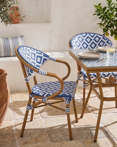Mediterranes Bistro-Set mit blau-weiß geflochtenen Gartenstühlen und Gestell in Holzoptik auf einer Steinterrasse.