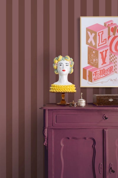 Vliestapete mit vertikalen Streifen in Braun und Rostrot hinter einer lila Kommode mit dekorativer Büste und gerahmtem Bild.