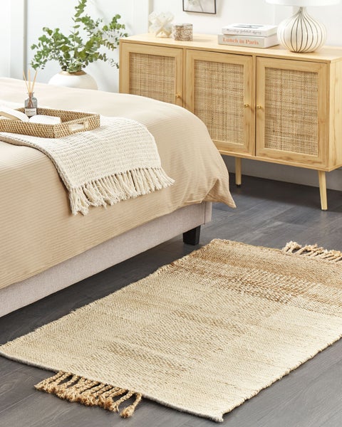 Helles Schlafzimmer mit einem Jutesteppich mit Fransen auf dunklem Boden, einem Bett mit beigen Textilien und einer Holzkommode mit Rattantüren.