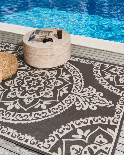 Heller Outdoor-Teppich mit ornamentalem Mandala-Muster auf einer strukturierten Poolterrasse aus Holz mit rundem Steintisch und Pouf aus Jute.