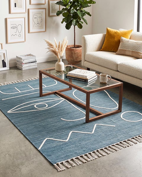 Wohnzimmer mit einem rechteckigen Teppich mit abstraktem Gesichtsmotiv und Fransen, der vor einem modernen Sofa und einem Glastisch liegt.