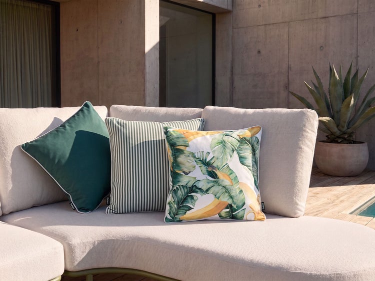 Drei quadratische Zierkissen mit unterschiedlichen Mustern auf einem hellen Outdoor-Sofa vor moderner Betonarchitektur und Agavenpflanze.