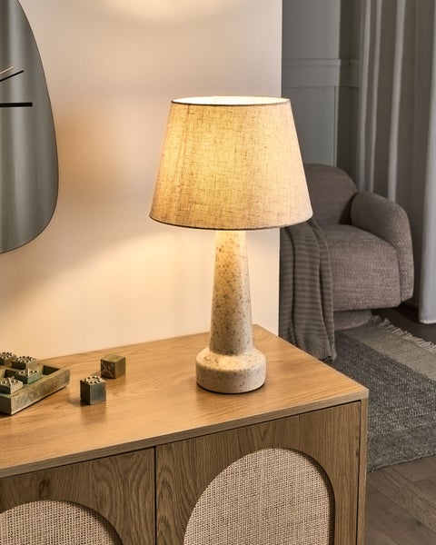 Tischleuchte mit gesprenkeltem Keramikfuß und Leinenschirm auf einem Sideboard aus Holz in einem wohnlich eingerichteten Zimmer.