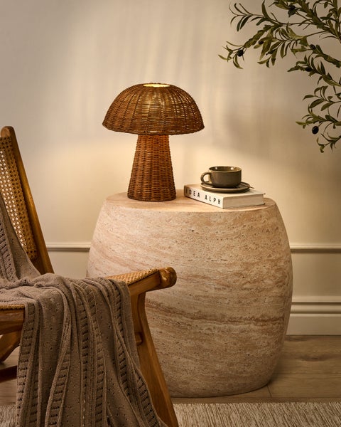 Dekorative Szene mit Rattanstischlampe, Buch und Tasse auf einem Steintisch neben einem Rattansessel mit Decke.