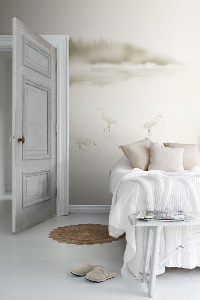 Helles Schlafzimmer mit Bett, Dekokissen, Decke, Beistelltisch, Zeitschriften und einer Tapete mit Reihermotiven