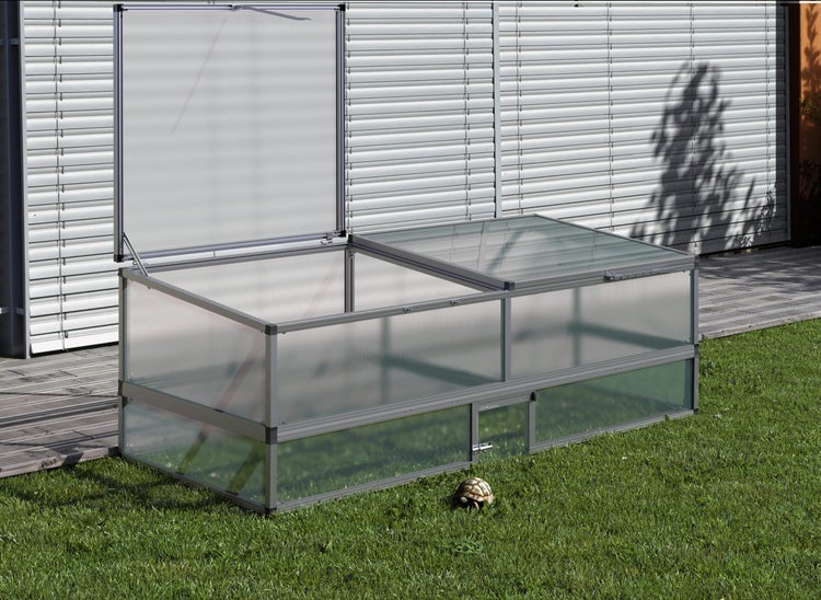 Schildkrötenhaus aus Aluminium und Doppelstegplatten auf einer Rasenfläche mit einem geöffneten Deckel, einer kleinen Tür und einer Schildkröte.