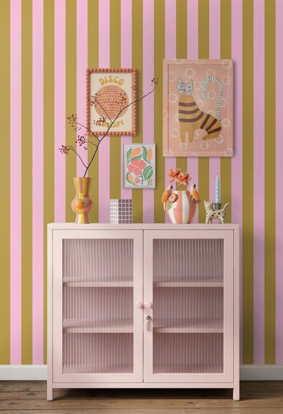 Innenraum mit rosa Vitrine vor pink-oliv gestreifter Tapete, dekoriert mit verschiedenen Vasen und gerahmten Kunstdrucken.