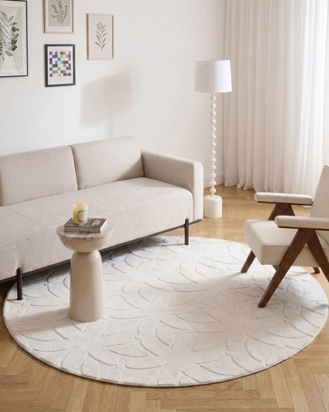 Helles Wohnzimmer mit Sofa, Sessel, rundem Teppich mit floralem Muster, Beistelltisch, Stehlampe und Wandbildern auf Holzboden.