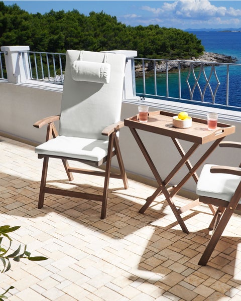 Hölzerne Gartenmöbel auf einer hellen Terrasse mit Meerblick, bestehend aus einem Klappsessel mit weißen Polstern und einem Beistelltisch.