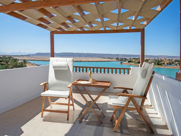 Holz-Gartenstühle mit hellen Polstern und ein hölzerner Klapptisch auf einem hellen Balkon mit Meerblick unter einer Pergola.