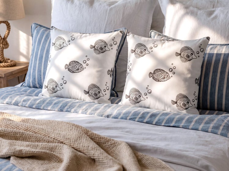 Helle Schlafzimmer-Szene mit Dekokissen mit Fischmotiven und blau gestreiften Kissen auf einem Bett mit passender Bettwäsche.