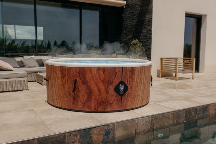 Runder aufblasbarer Whirlpool in Holzoptik auf einer modernen Terrasse neben einem Pool. Coco Spa Logo auf der Außenseite.