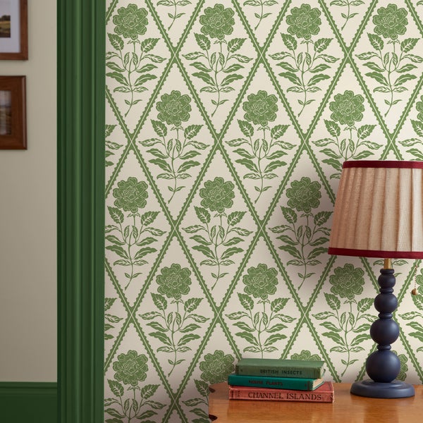 Vliestapete mit Rautenmuster und floralem Motiv an einer Wand, dekoriert mit einer Tischlampe und Büchern auf einer Holzkommode.