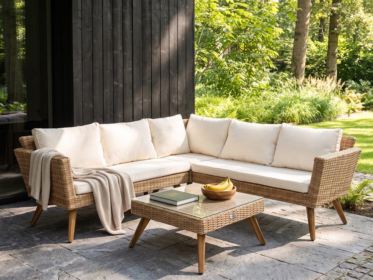 Eck-Gartenlounge-Set aus Polyrattan mit hellen Polstern und Couchtisch mit Glasplatte auf einer Steinterrasse vor einer dunklen Holzwand.