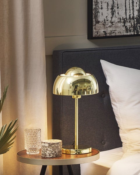 Goldfarbene Tischleuchte in Pilzform auf einem hölzernen Nachttisch neben einem Bett mit dunkelgrauem Kopfteil und weißem Kissen.