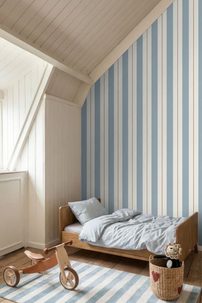 Gestreifte Tapete in einem Kinderzimmer mit Holzbett unter einer Dachschräge und einem Holz-Laufrad auf einem Teppich.