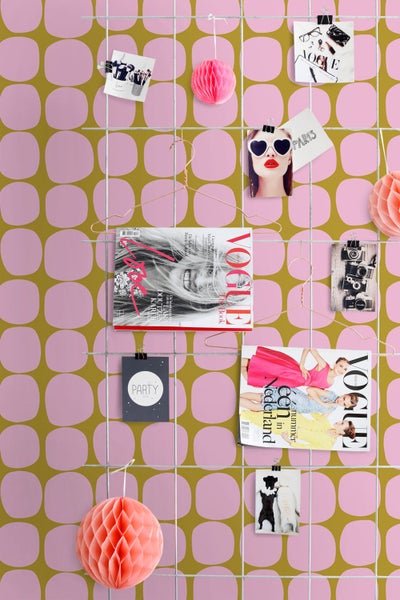 Retro-Mustertapete in Pink und Ocker mit einem Metallgitter als Pinnwand, bestückt mit Zeitschriften, Fotos und Papierlampions.