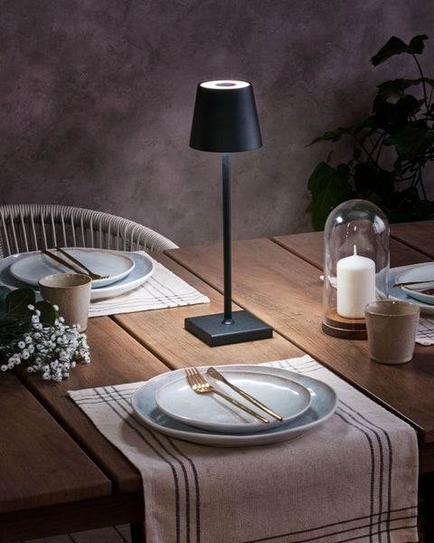 Moderne schwarze Tischleuchte auf einem Holztisch mit Keramikgeschirr, Goldbesteck und Kerze in einem stimmungsvoll beleuchteten Ambiente.