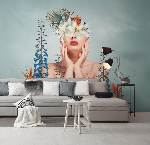 Wohnzimmer mit Fototapete eines Frauenporträts mit Lilien und blauen Blumen. Graues Ecksofa mit Kissen, weißer Tisch und schwarze Stehlampe.