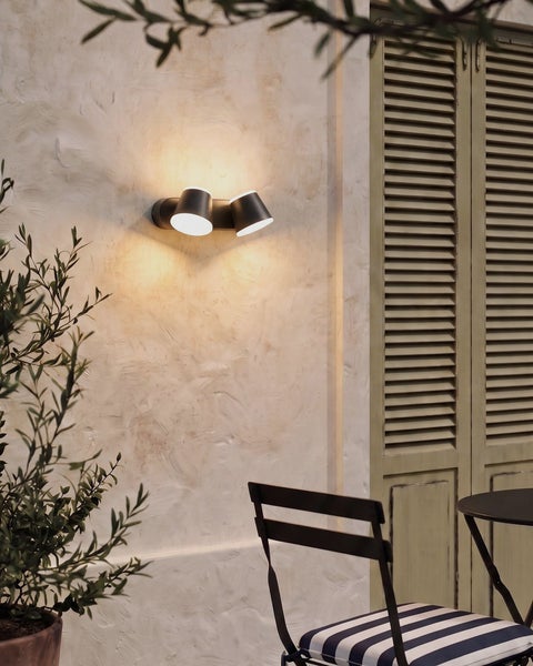 Zweiflammige Außenwandleuchte mit zwei verstellbaren Spots, warm leuchtend an einer mediterranen Hauswand montiert.