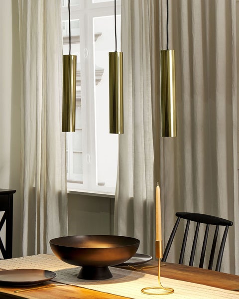 Drei goldfarbene, zylindrische Pendelleuchten aus Metall hängen über einem Holztisch mit Geschirr in einem hellen Raum mit Vorhängen.