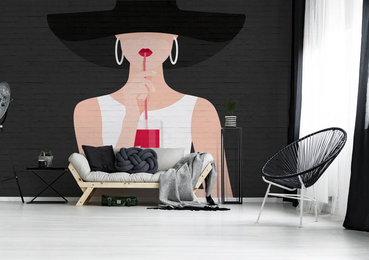 Fototapete mit Illustration einer Frau mit Hut auf einer Wand in Backsteinoptik hinter einem Sofa aus hellem Holz und einem schwarzen Stuhl.