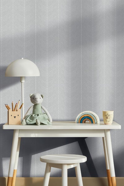 Kinderzimmer mit weißem Schreibtisch, Hocker und Lampe vor grauer Tapete mit vertikalem Fischgrätmuster.