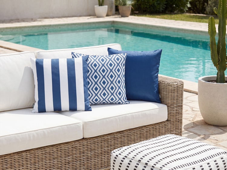 Drei dekorative Gartenkissen auf einem Rattan Sofa mit Pool im Hintergrund.