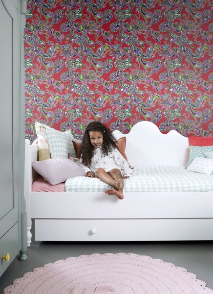 Mädchen sitzt auf einem Bett vor einer gemusterten Tapete in einem Kinderzimmer.