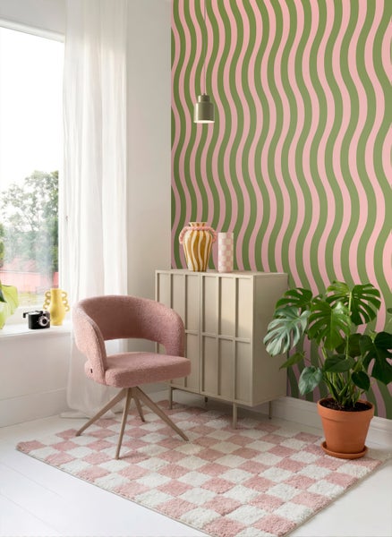 Modernes helles Zimmer mit grün-rosa Wellentapete, rosa Polstersessel, beigem Sideboard, kariertem Teppich und großer Monstera-Zimmerpflanze.