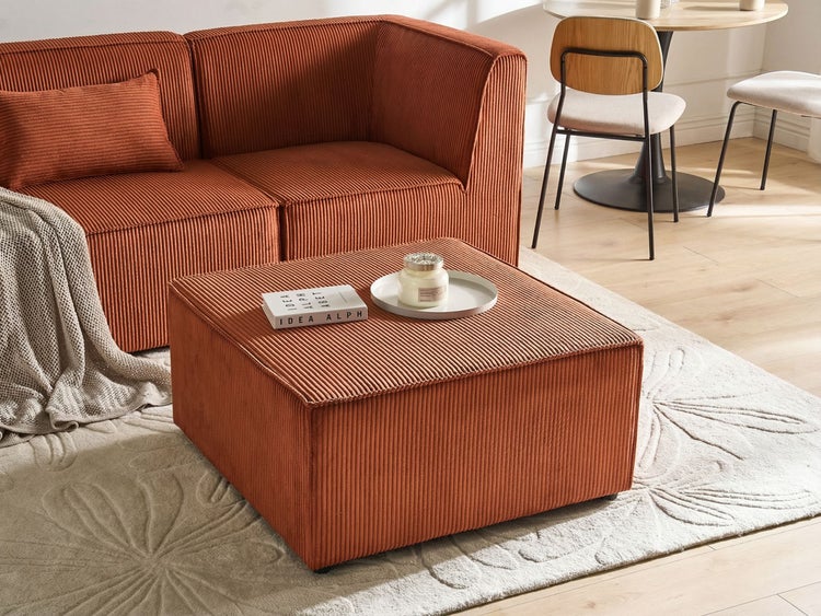 Hell eingerichtetes Wohnzimmer mit Ecksofa und quadratischem Hocker aus orangefarbenem Cord, gemustertem Teppich und Essgruppe im Hintergrund.