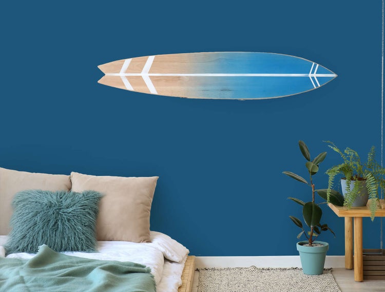 Wanddekoration in Surfbrett-Form an einer dunkelblauen Wand über einem Bett mit Pflanzen im modernen Schlafzimmer.