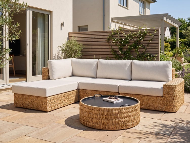 Gartenlounge-Set mit einem Ecksofa in Flechtoptik und einem runden Couchtisch auf einer hellen Terrasse.
