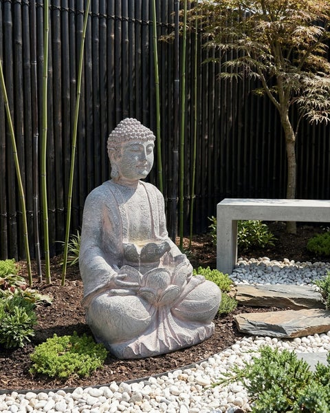Buddha Gartenbrunnen in Steinoptik im Zen Garten mit Bambuszaun, hellen Kieselsteinen und einer Betonbank.