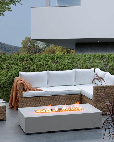 Moderne Terrassengestaltung mit rechteckigem Gas-Feuertisch, einer Lounge-Eckcouch aus Polyrattan mit weißen Kissen und einer grünen Hecke.