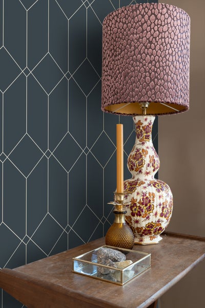 Nahaufnahme eines Tisches mit Lampe, Kerze und einer Schale mit Steinen vor einer Wand mit geometrischer Tapete.