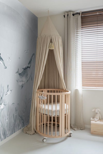 Ein Kinderzimmer mit einem runden Babybett, einem Baldachin, einer Tapete mit Unterwassermotiven und einem Fenster mit Jalousien.