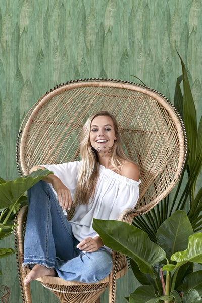 Frau sitzt in einem Rattansessel vor einer grünen Tapete mit Blattmuster
