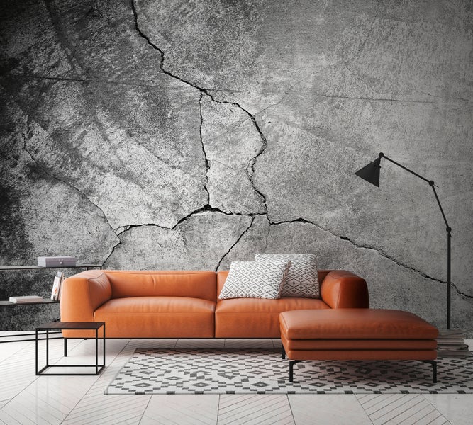 Modernes Wohnzimmer mit Fototapete in Betonoptik mit Rissen, orangefarbenem Ledersofa, Hocker und geometrischem Teppich auf hellem Holzboden.