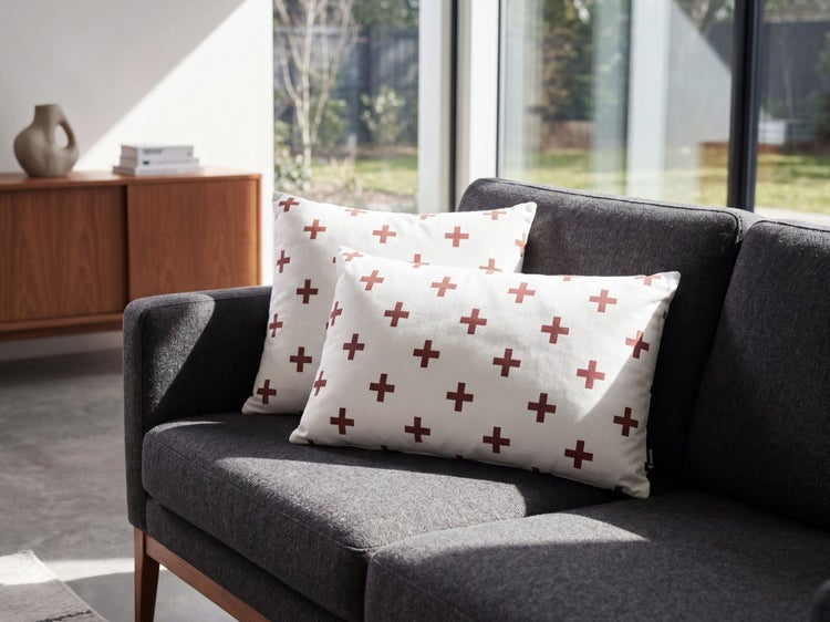 Zwei weiße Dekokissen mit braunrotem Plus-Muster auf einem dunkelgrauen Sofa in einem hellen, modernen Wohnzimmer.