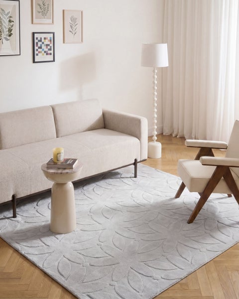 Wohnzimmer mit beigem Sofa, Sessel, skulpturalem Beistelltisch, Stehlampe und einem gemusterten Teppich mit floralem Relief auf Parkettboden.