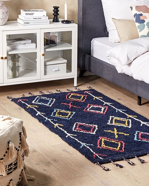 Dunkelblauer Teppich mit bunten geometrischen Mustern und Quasten auf Holzfußboden im Schlafzimmer neben grauem Bett und weißem Sideboard.