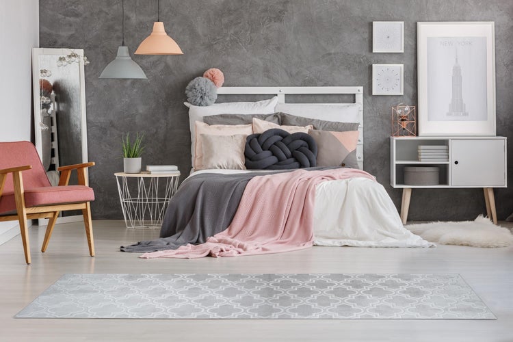 Modernes Schlafzimmer mit grau strukturierter Wand, weißem Bett, rosa Sessel, gemustertem Teppich und Pendelleuchten in Grau und Apricot.