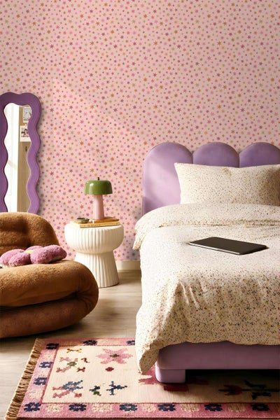 Kinderschlafzimmer mit Tapete mit Sternenmuster, lila gepolstertem Bett, geschwungenem Spiegel, Sessel und gemustertem Teppich.