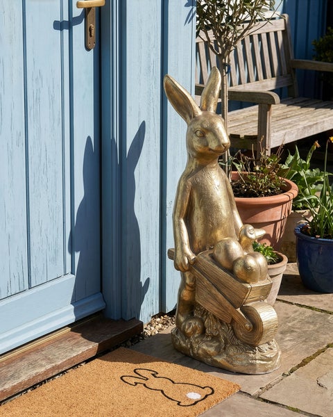 Goldene Gartenfigur Hase mit Schubkarre und Eiern vor einer blauen Tür mit Fußmatte und Pflanzen.