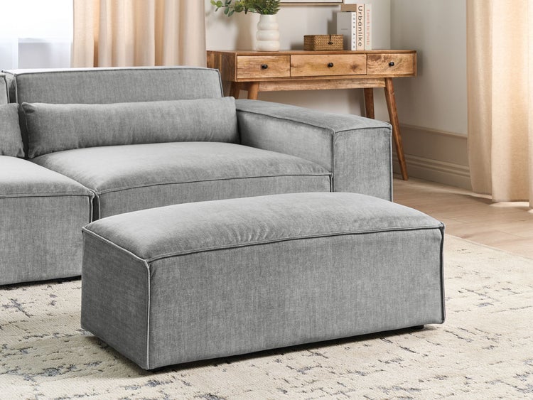 Gepolsterter grauer Hocker vor einem Stoffsofa in einem hellen Wohnzimmer mit Teppich und Konsolentisch aus Holz.