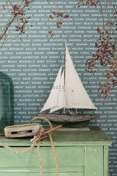 Dekorative Segelboot-Miniatur auf einem grünen Schrank mit Seil und Flaschenzug vor einer Tapete mit nautischem Motiv